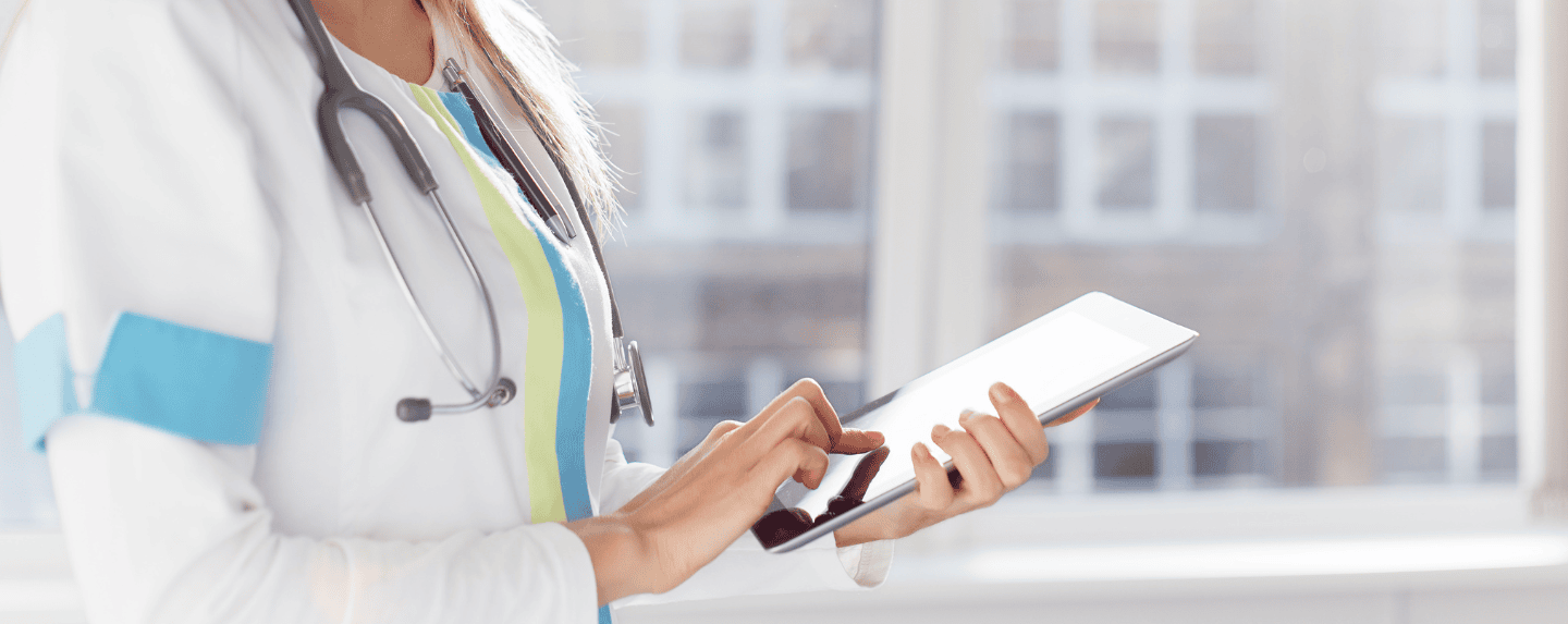 PelicanSoft - A medical professional wearing a white coat and stethoscope uses a tablet equipped with custom healthcare software in a well-lit room with large windows, enhancing patient care.